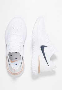 White athletic shoes with mesh material, featuring a navy swoosh, gold accents, and a textured sole. Includes a cushioned insole with hexagonal patterns.
