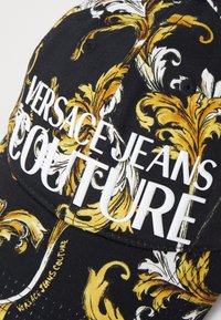 Črna kapa s zlatim in belim okrasnim vzorcem ter izbočeno belim besedilom, ki pravi "VERSACE JEANS COUTURE." Teksturirana površina tkanine.