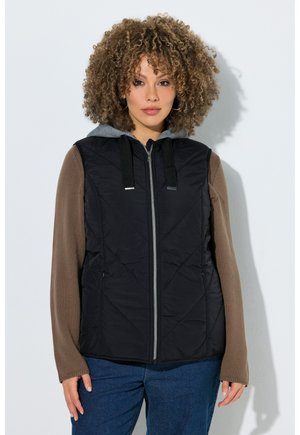 Vrouw met krullend haar die een zwart vest met ritssluiting draagt over een bruin shirt met lange mouwen en een blauwe spijkerbroek, staand tegen een witte muur.