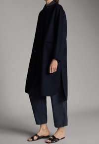 Cappotto lungo blu navy con taglio dritto, chiusura a bottoni, due tasche laterali e spacchi laterali, abbinato a pantaloni scuri e sandali neri.