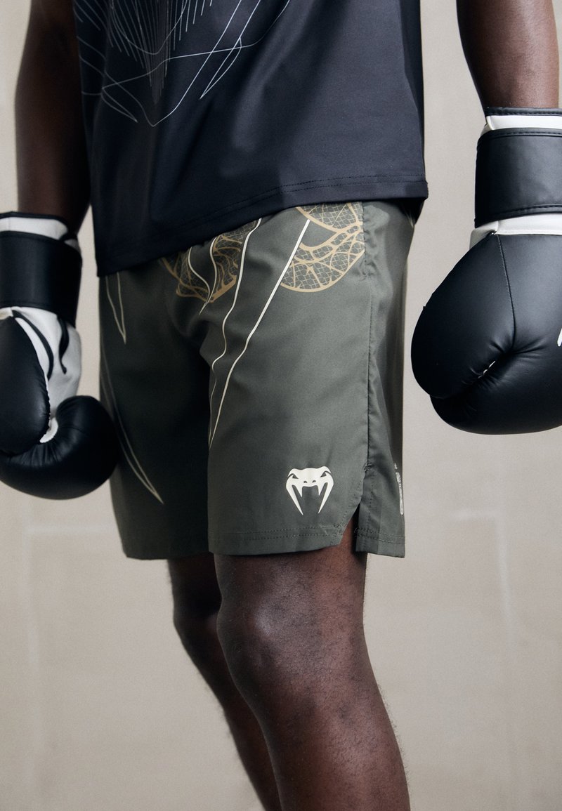 Pantalones de boxeo en verde oliva, con un patrón de telaraña dorada y un logo blanco. Tela suave con aberturas laterales para mayor movilidad. Guantes negros puestos.