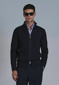 Marineblauer, gerippter Reißverschluss-Pullover mit hohem Kragen, silberner Reißverschluss und langen Ärmeln, getragen über einem hellblauen Hemd mit Kragen. Glatte Textur.