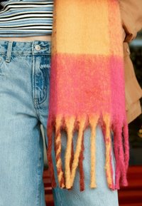 Mehrfarbiger Schal aus orangefarbenem, pinkem und gelbem Wolle, mit Fransenenden und einer weichen Textur, neben hellblauen Jeans drapiert.