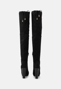 Botas negras por encima de la rodilla hechas de ante. Presentan un tacón alto, un cordón ajustable en la parte superior y cremalleras en los laterales. Textura suave.