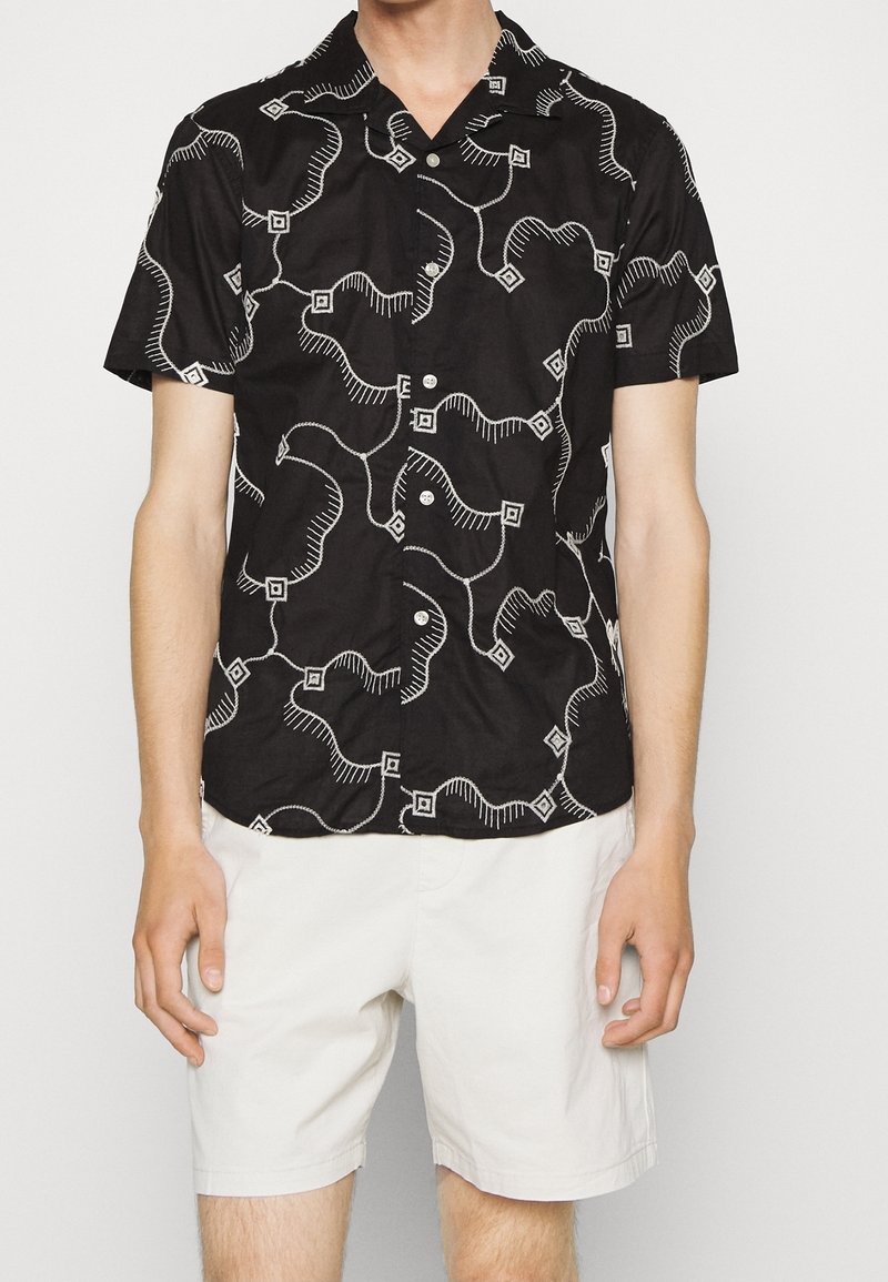 Homme portant une chemise noire à manches courtes à boutons avec un motif abstrait de lignes blanches et un short blanc, debout devant un fond uni.