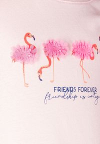 Jasnoróżowa koszulka z czterema flamingami ozdobionymi różowymi kwiatowymi akcentami, teksturowanymi płatkami i niebieskim napisem „Przyjaciele na zawsze”.