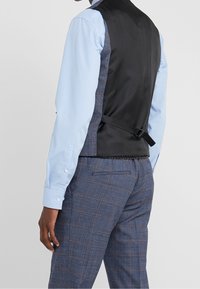 Man wearing blue checkered pants, light blue dress shirt, and adjustable black-back vest, shown from the back against a plain background.