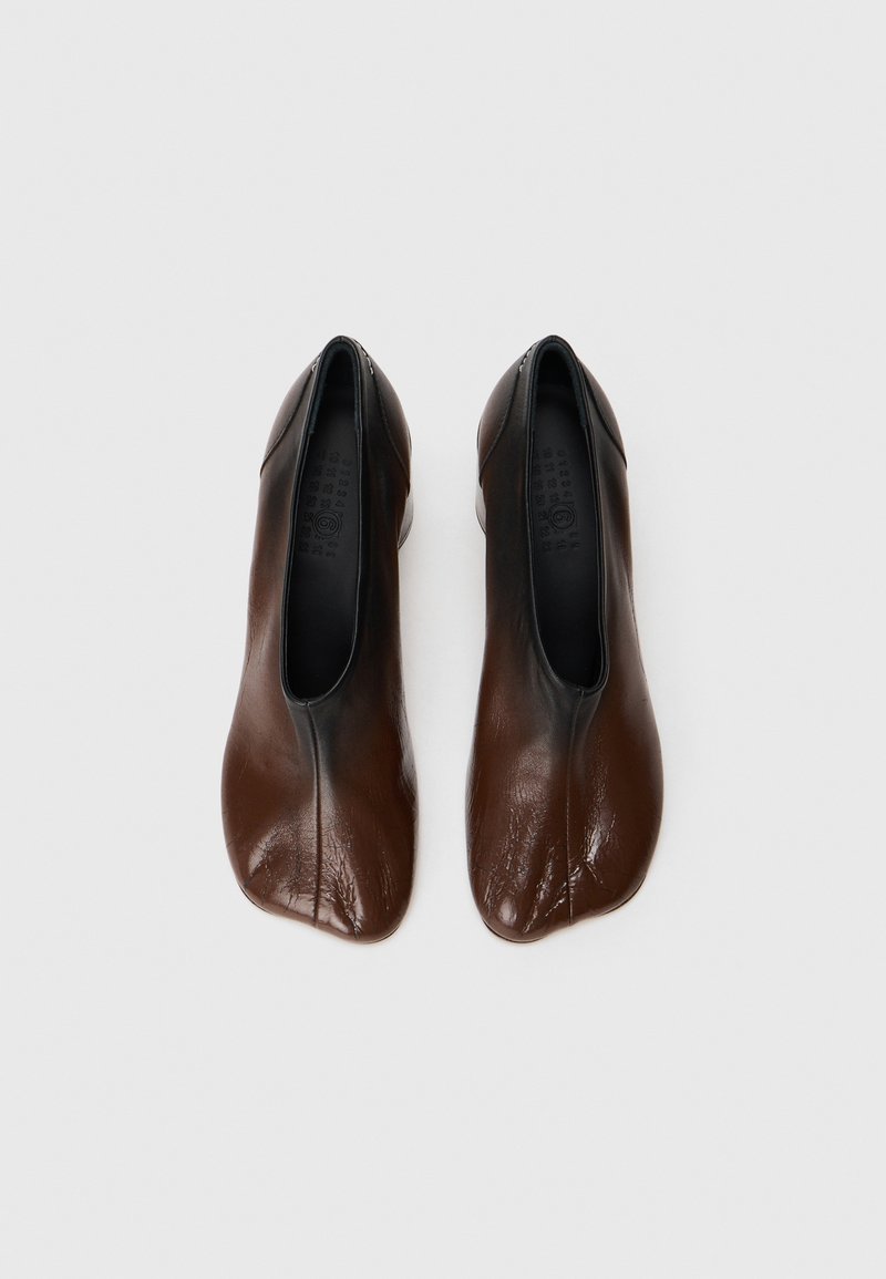 Un paio di scarpe in pelle marrone senza lacci con una particolare punta rotonda e irregolare su uno sfondo bianco uniforme.