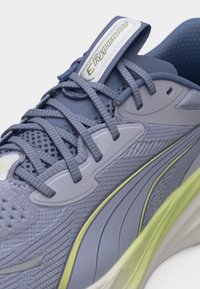 Εγχώρια λήψη ενός γκρι και κίτρινου παπουτσιού τρεξίματος με υφασμάτινο επάνω μέρος με υφή, μαξιλαρωμένη γλώσσα και κορδόνια με σχέδιο δεμένα σε απλό κόμπο.