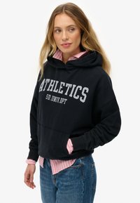 Mujer con cabello rubio largo que lleva una sudadera negra con la palabra "ATHLETICS" sobre una camisa de rayas rosa y jeans azules, mirando ligeramente hacia su izquierda.