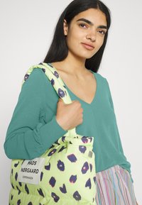 Camisola verde de mangas longas com decote em V, combinada com uma bolsa tote almofadada em verde claro com manchas roxas e azul-marinho e um detalhe de etiqueta branca.