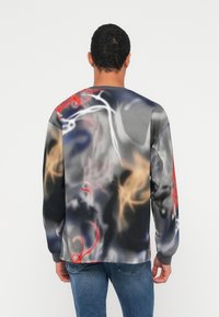 Άνδρας που φορά γκρι μακρυμάνικο πουκάμισο με αφηρημένα κόκκινα, λευκά, μαύρα και μπεζ σχέδια, συνδυασμένο με μπλε τζιν, γυρισμένος προς τα πίσω σε απλό φόντο.