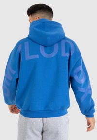 Haut à capuche bleu en coton, avec un grand texte graphique violet au dos. Dispose d'une capuche et de poignets côtelés. Design à coupe régulière.