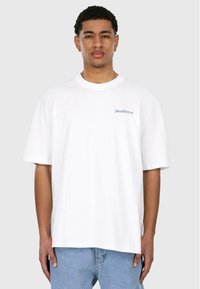 White cotton t-shirt with short sleeves, round neckline, and blue logo detail on the chest. Worn with light blue denim jeans.