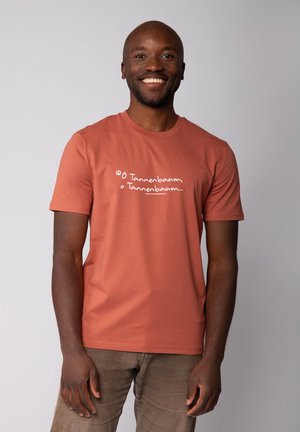 Glunderende man in een roestkleurig T-shirt met handgeschreven tekst "O Tannenbaum, o Tannenbaum...", staande tegen een effen lichte achtergrond.