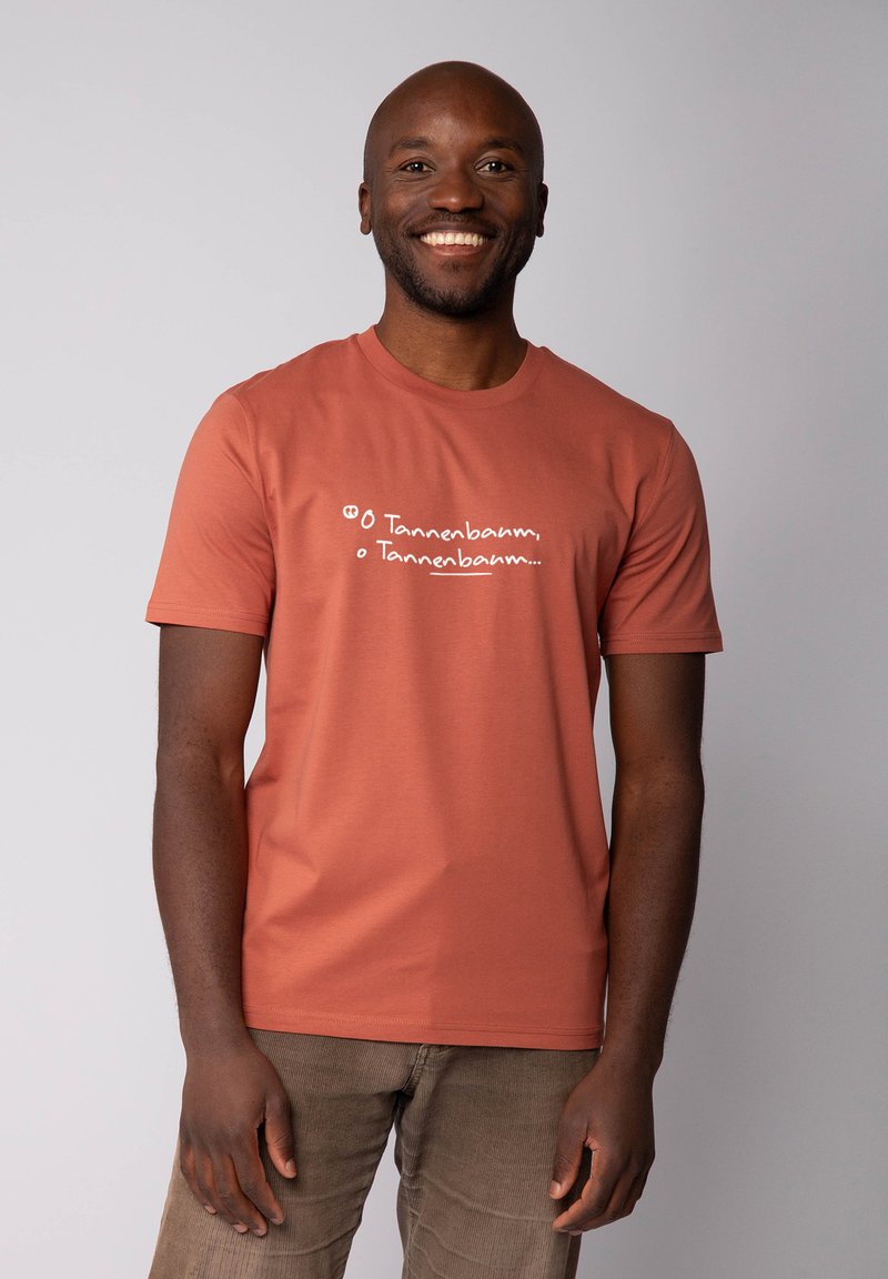 Homme souriant portant un T-shirt couleur rouille avec le texte manuscrit "O Tannenbaum, o Tannenbaum...", se tenant devant un fond clair.