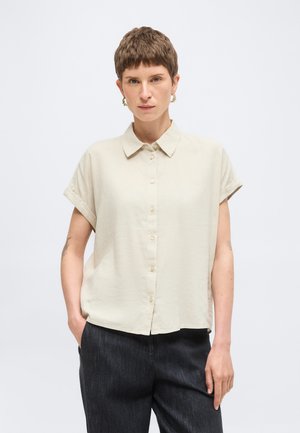 Osoba o krótkich włosach ubrana w beżową koszulę na krótki rękaw z guzikami i ciemne spodnie, stojąca z jedną ręką w kieszeni.