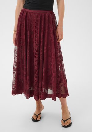 Femme portant une longue jupe en dentelle bordeaux avec des motifs floraux et des sandales noires, debout devant un fond clair uni.