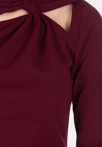 Indumento a maniche lunghe color bordeaux con dettaglio ad nodo attorcigliato vicino alla spalla e un ritaglio, realizzato in tessuto liscio ed elastico.