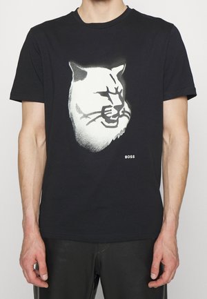 T-shirt noir à manches courtes avec un visage de chat stylisé en blanc et un petit texte "BOSS" sur le devant, porté par une personne aux bras détendus.