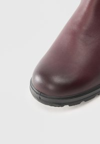 Bota de cuero burdeos con punta redondeada, parte superior texturizada y suela de goma negra. Un detalle de costura a lo largo de la zona del dedo aporta interés visual.