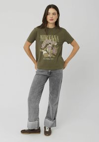 T-shirt en coton vert olive avec un graphique floral Nirvana en doré et violet. Associé à un jean large gris et des baskets brunes.