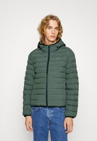 Zelená puffer bunda s kapucí, má zipové zapínání a přišitý vzor. Logo Lacoste na levé hrudi, v kombinaci s modrými džíny.