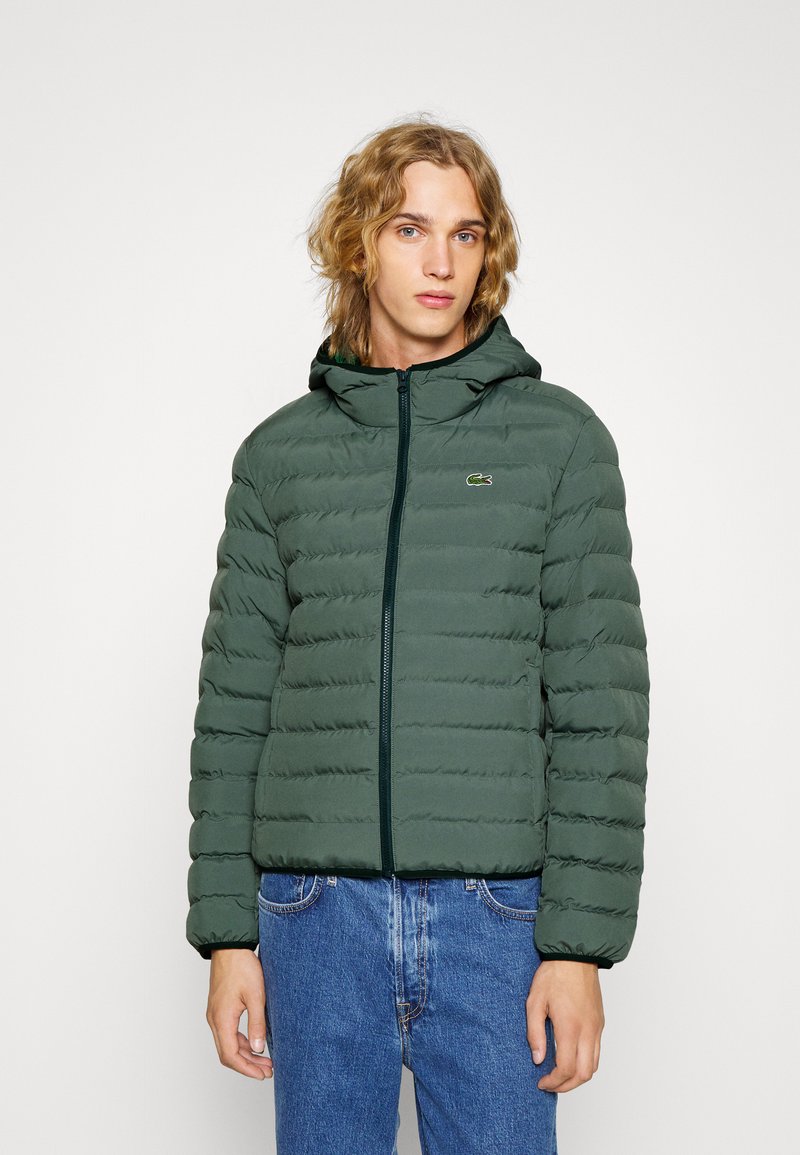 Zelená puffer bunda s kapucí, má zipové zapínání a přišitý vzor. Logo Lacoste na levé hrudi, v kombinaci s modrými džíny.