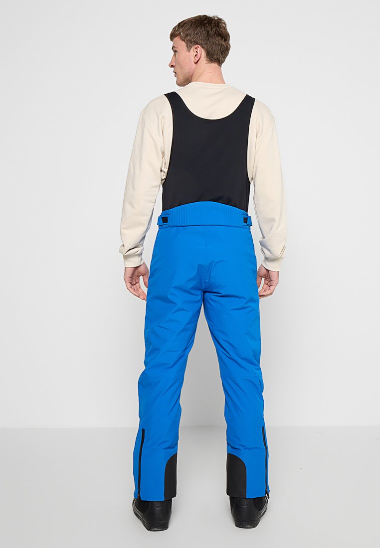 Mężczyzna zwrócony tyłem, ubrany w czarny bezrękawnik oraz jaskrawo niebieskie spodnie narciarskie z regulowanym pasem i czarnymi butami.