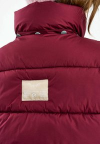 Bordó puffert dzseki hátul, bézs színű téglalap alakú cimkével, és világosbarna haj a gallér belsejébe tűrve.