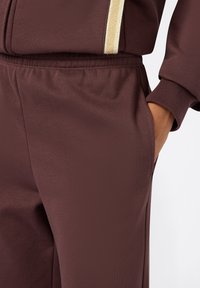 Bordeauxkleurige sweatpants met een gladde textuur, voorzien van een elastische tailleband, zijzakken en een subtiele gouden accenten langs de rits.