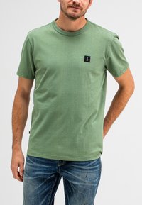 Groene katoenen T-shirt met korte mouwen, ronde hals en zwart logo-embleem. Draag je met blauwe denim jeans, met een relaxed fit en een lichte slijtage.