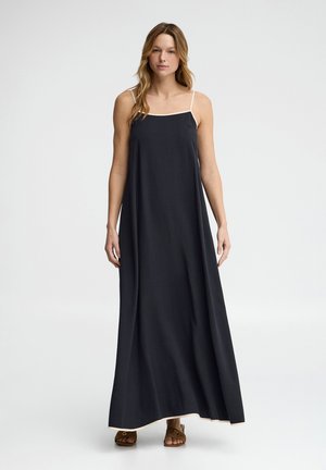 Kobieta ubrana w długą czarną sukienkę bez rękawów z białym wykończeniem i brązowe sandały z odkrytymi palcami, stojąca na jednolitym białym tle.