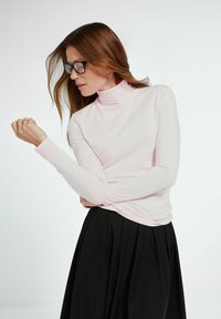 Kobieta z długimi brązowymi włosami, nosząca czarne okulary, jasnoróżowy golf oraz czarną spódnicę, patrzy na lewy nadgarstek.