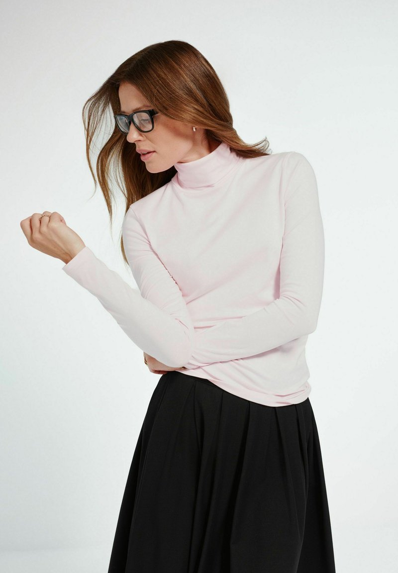 Kobieta z długimi brązowymi włosami, nosząca czarne okulary, jasnoróżowy golf oraz czarną spódnicę, patrzy na lewy nadgarstek.