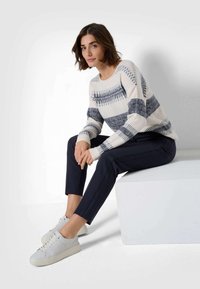 Maglione chiaro con strisce navy e motivi geometrici, abbinato a pantaloni navy e sneaker bianche, seduto su un blocco bianco.