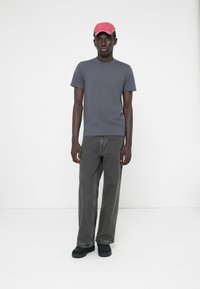 Un tricou gri, blugi întunecați largi și pantofi negri. O șapcă roz cu accente albe completează ținuta. Fundal simplu.