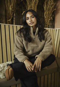 Beżowy polarowy golf z ćwierć zamkiem, z teksturowanym wykończeniem i elastycznymi mankietami, zestawiony z czarnymi spodniami i beżowymi botkami za kostkę.