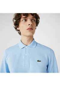 Lyseblå polo skjorte med struktureret stof, tre knaplukninger og grønt Lacoste-logo på venstre bryst. Korte ærmer.