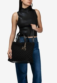 Borsa a tote nera in tessuto intrecciato con dettagli in pelle, dotata di un ciondolo decorativo dorato. Forma strutturata, ampio interno e manici superiori.