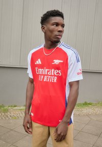 Maillot de football rouge avec des manches blanches, arborant le texte "Emirates FLY BETTER". Accents bleus et tissu texturé. Modèle portant un pantalon kaki.