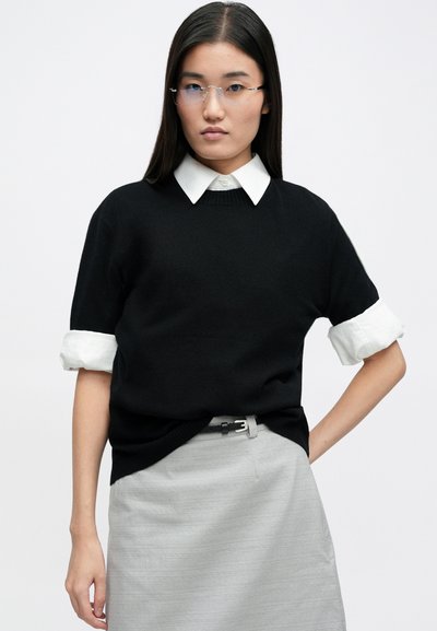 Black short-sleeve sweater over a white collared shirt, with the shirt cuffs rolled. Accented with a light gray skirt and a thin belt.