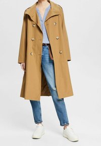 Trench coat marrone chiaro con ampi revers, bottoni doppi e vestibilità comoda. Indossato con jeans blu strappati e scarpe da ginnastica bianche.