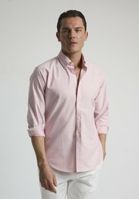 Camisa de botón rosa claro con cuello, mangas largas arremangadas en los puños, combinada con pantalones blancos. Tejido suave, con un diseño minimalista.