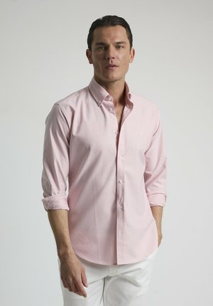 Camisa de botón rosa claro con cuello, mangas largas arremangadas en los puños, combinada con pantalones blancos. Tejido suave, con un diseño minimalista.