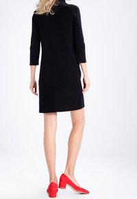 Robe noire à manches trois-quarts, texture lisse, longueur genou. Assortie à des escarpins en daim rouge, avec une forme effilée et un talon en bois.