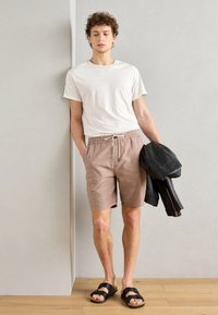 T-shirt à col rond blanc, shorts beige clair avec cordon, et sandales noires. Tenant une veste en cuir noire. Fond et sol neutres.