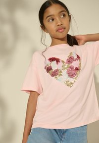 Rosa T-Shirt mit kurzen Ärmeln und großem Herzdesign aus Pailletten in Rot-, Pink- und Goldtönen. Lässig geschnitten, kombiniert mit blauer Jeans.