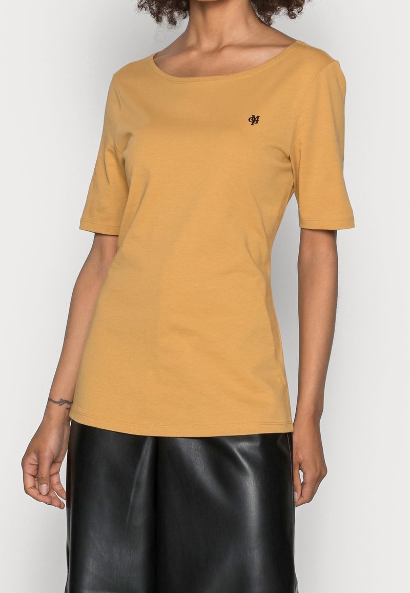 Musztardowa, krótko rękawowa koszulka z miękkiej bawełny, z dekoltem w łódkę i małym czarnym logo na lewej piersi. Połączona ze skórzaną spódnicą.
