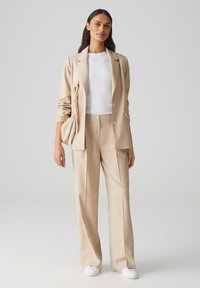 Beige op maat gemaakte blazer en broek met een witte top. Gladde stof, klassieke revers en subtiele zakdetails. Witte sneakers als schoeisel.
